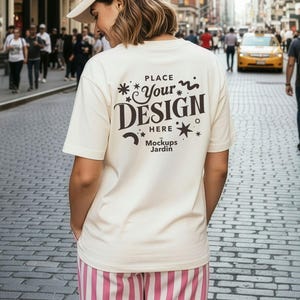 Puede incluir: Una persona con una camiseta color crema con el texto "PLACE YOUR DESIGN HERE" en una fuente decorativa. La persona lleva una gorra de béisbol beige y pantalones a rayas rosas y blancas. El fondo es una calle de la ciudad con peatones y edificios.