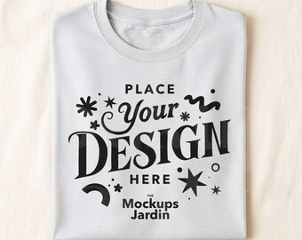 May include: A folded light gray t-shirt with a crew neck. The shirt features the text "PLACE Your DESIGN HERE" in a black, decorative font, along with the text "The Mockups Jardin".