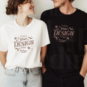 Op de afbeelding: Twee mensen dragen witte en zwarte T-shirts met de tekst "PLACE YOUR DESIGN HERE Mockups Jardin" gedrukt op de voorkant. De mensen staan voor een witte bakstenen muur.