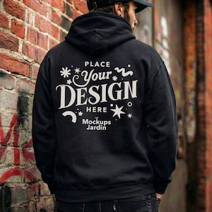 May include: Black hooded sweatshirt with the text "PLACE Your DESIGN HERE Mockups Jardin" in white. The sweatshirt is shown against a brick wall background. The person wearing the sweatshirt is facing away from the camera.