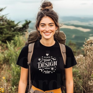 May include: A woman wearing a black t-shirt with the text "PLACE YOUR DESIGN HERE" and a brown backpack. She is standing in a green grassy area with a mountain in the background.