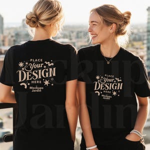 Puede incluir: Dos personas con camisetas negras con el texto "PLACE YOUR DESIGN HERE Mockups Jardin" en blanco. Las camisetas son de manga corta. El fondo muestra un paisaje urbano con edificios altos y un cielo brillante.
