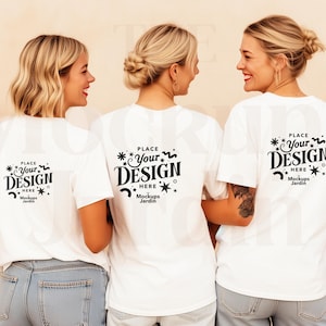 May include: Three women wearing white t-shirts and blue jeans. The t-shirts have a black design on the back that says "PLACE Your DESIGN HERE Mockups Jardin". The women are smiling and standing together.