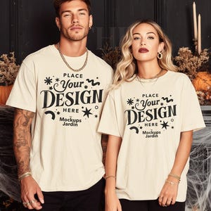May include: Two people wearing cream-colored t-shirts with the text "PLACE Your DESIGN HERE Mockups Jardin" in black. The shirts are paired with black pants. The background includes pumpkins, candles, and decorative elements, suggesting a Halloween theme.