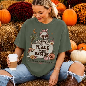 May include: An olive green t-shirt featuring a graphic of a skeleton wearing a checkered headscarf and heart-shaped sunglasses, leaning on a sign that reads PLACE YOUR DESIGN. The design includes a ghost, palm tree, and pumpkins.