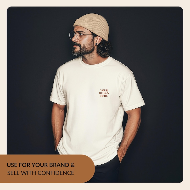 Może przedstawiać: Kremowy t-shirt z kr&oacute;tkim rękawem i okrągłym dekoltem, z napisem "YOUR DESIGN HERE" wydrukowanym ciemnobrązową czcionką szeryfową na lewej piersi. Koszulka jest noszona z beżową czapką z prążkowanej dzianiny i okularami w cienkich oprawkach.