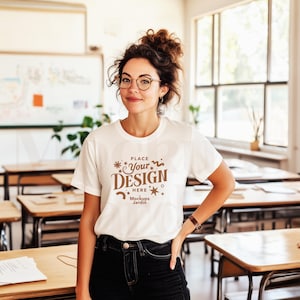 A stylish female teacher wearing a white Bella Canvas 3001 t-shirt stands confidently in a bright, modern classroom. With her curly hair in a loose updo, trendy glasses, and a warm smile, she embodies a professional yet relaxed look. The neatly arranged desks, large windows, and natural light create an inviting academic setting, making this mockup ideal for showcasing casual, work-appropriate apparel for educators and creative professionals.