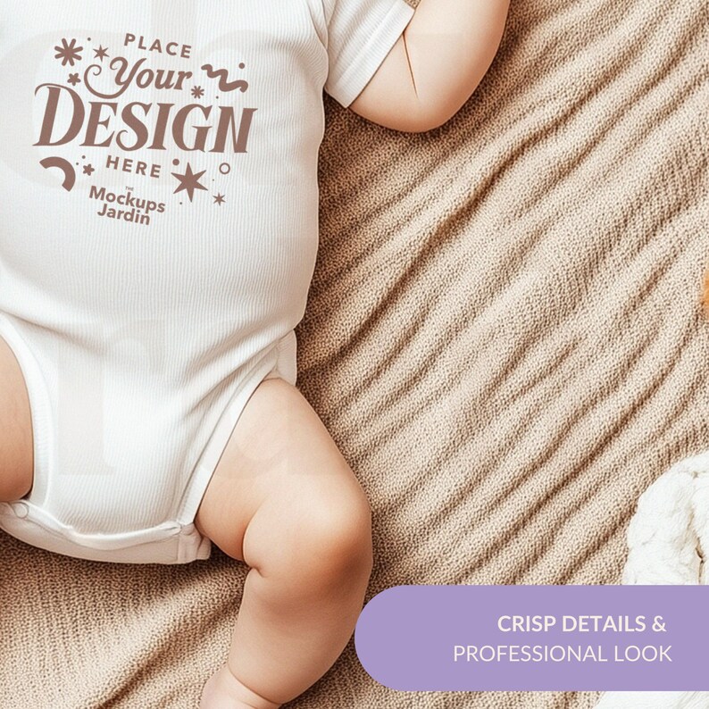 May include: White baby onesie with the text "Place Your Design Here" and "Mockups Jardin" printed on the front. The onesie is lying on a beige blanket.