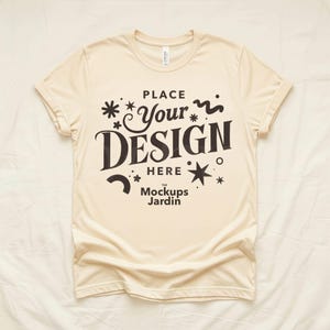 May include: A cream-coloured t-shirt with rolled-up sleeves. The shirt displays the text "PLACE Your DESIGN HERE" in a bold, dark font, with decorative star and swirl elements. The words "the Mockups Jardin" are at the bottom.