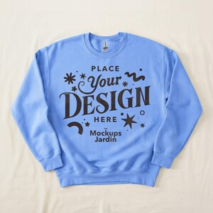 Op de afbeelding: Een lichtblauw sweatshirt met ronde hals en de tekst "PLACE Your DESIGN HERE" in zwart, samen met decoratieve ster- en krulafbeeldingen. Het Mockups Jardin-logo staat onderaan. Het sweatshirt wordt gepresenteerd op een crèmekleurig oppervlak.