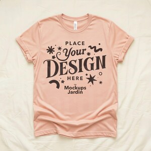 Puede incluir: Una camiseta rosa claro con las mangas enrolladas. La camiseta presenta el texto "PLACE Your DESIGN HERE" en marrón oscuro, junto con elementos decorativos de estrellas y remolinos. También está presente el texto "The Mockups Jardin".