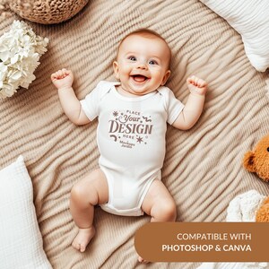 May include: White baby onesie with short sleeves and a brown text overlay that says "Place Your Design Here Mockups Jardin". The onesie is on a brown blanket with a white pillow and a white flower bouquet.