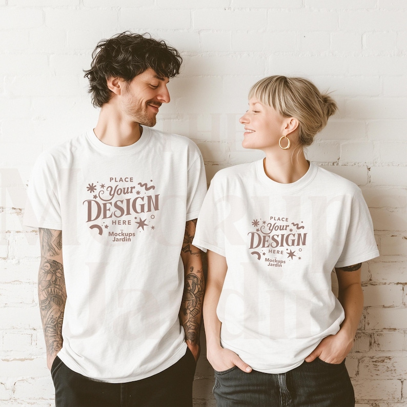 May include: Two people wearing white t-shirts with the text "PLACE *Your DESIGN HERE Mockups Jardin" printed on the front. The people are standing in front of a white brick wall.
