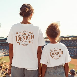 以下が含まれることがあります： 茶色の「PLACE Your DESIGN HERE Mockups Jardin」の文字が入った白いTシャツが2枚。2人の人が着ており、背中がカメラに向いています。野球場の外に立っています。