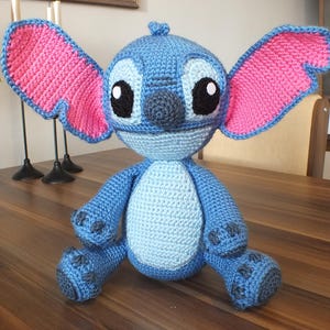 May include: A handmade blue crochet Stitch doll with large pink-lined ears, black eyes, and a light blue belly. The plush toy is sitting on a wooden table. A red poinsettia plant in a red pot is in the background.
