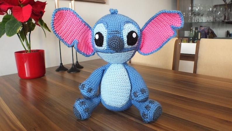 May include: A handmade blue crochet Stitch doll with large pink-lined ears, black eyes, and a light blue belly. The plush toy is sitting on a wooden table. A red poinsettia plant in a red pot is in the background.