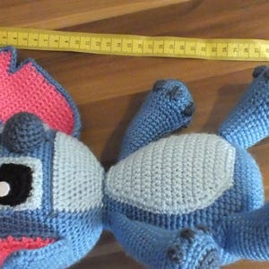 May include: A handmade blue crochet Stitch doll with pink inner ears and a gray belly. The doll is lying on a wooden surface with a yellow measuring tape. The doll's length is approximately 12 inches.
