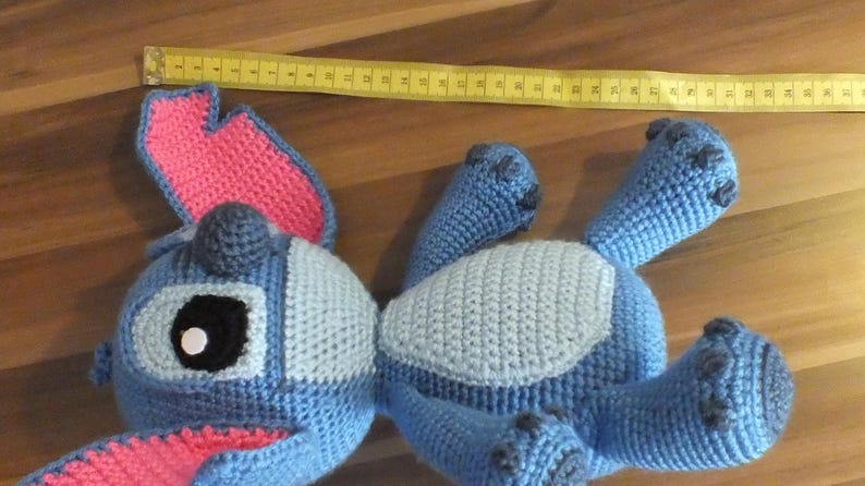 May include: A handmade blue crochet Stitch doll with pink inner ears and a gray belly. The doll is lying on a wooden surface with a yellow measuring tape. The doll's length is approximately 12 inches.