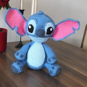 May include: A handmade blue and pink crochet Stitch doll. The doll has large ears with pink interiors, big black eyes, and a gray nose. It is sitting on a wooden surface. The doll is a character from the animated movie Lilo & Stitch.