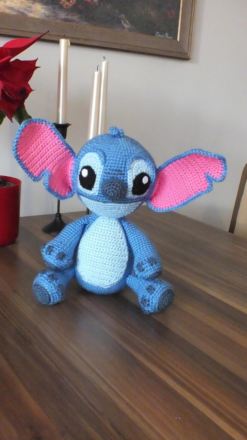 May include: A handmade blue and pink crochet Stitch doll. The doll has large ears with pink interiors, big black eyes, and a gray nose. It is sitting on a wooden surface. The doll is a character from the animated movie Lilo & Stitch.