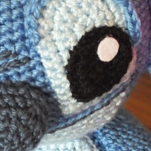 May include: Close-up of a crocheted blue and white character with a large black eye and a white pupil. The character has a dark gray nose and a pink ear. The crochet stitches are visible, creating a textured appearance.