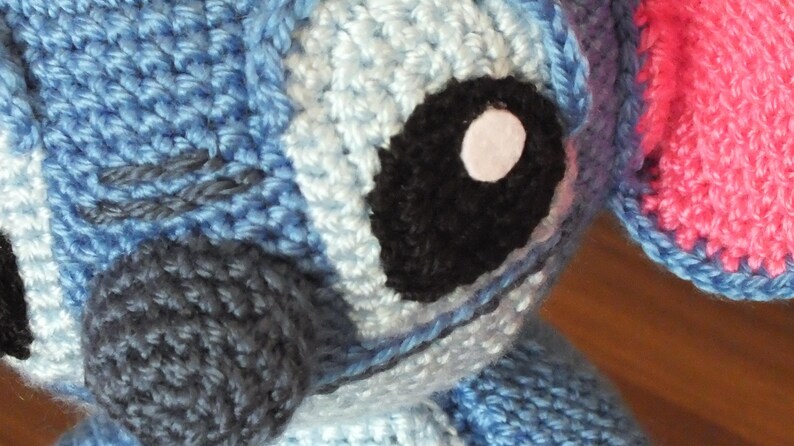 May include: Close-up of a crocheted blue and white character with a large black eye and a white pupil. The character has a dark gray nose and a pink ear. The crochet stitches are visible, creating a textured appearance.