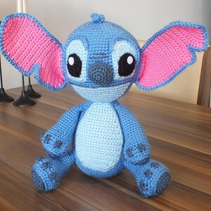May include: A handmade blue crochet Stitch doll with pink ears and black eyes. The plush toy is sitting on a wooden table. The doll is approximately 10 inches tall.