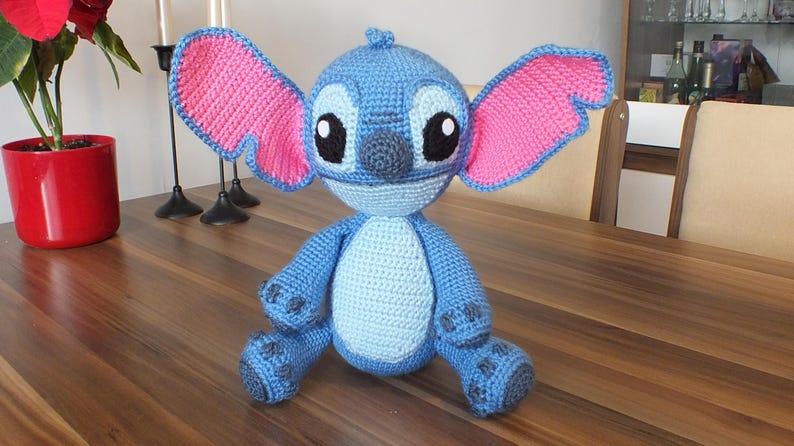 May include: A handmade blue crochet Stitch doll with pink ears and black eyes. The plush toy is sitting on a wooden table. The doll is approximately 10 inches tall.