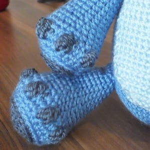 May include: Close-up of a crocheted blue stuffed animal's feet and lower body. The feet are a darker blue with gray accents, and the body is a lighter blue. The stitches are clearly visible, showing the handmade nature of the toy.