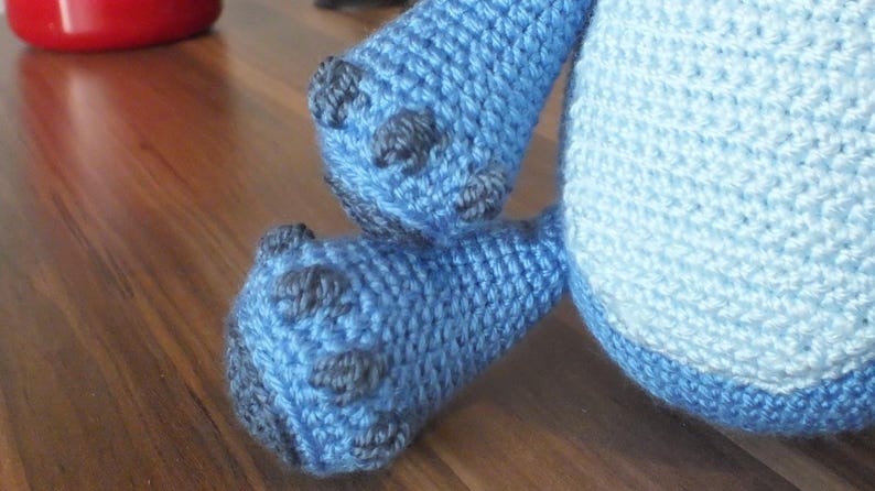 May include: Close-up of a crocheted blue stuffed animal's feet and lower body. The feet are a darker blue with gray accents, and the body is a lighter blue. The stitches are clearly visible, showing the handmade nature of the toy.