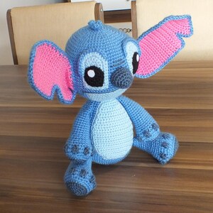 May include: A handmade blue crochet Stitch doll with pink inner ears, black eyes, and a gray nose. The plush toy is sitting on a wooden table. The doll's body is light blue, with darker blue accents on the feet and hands.