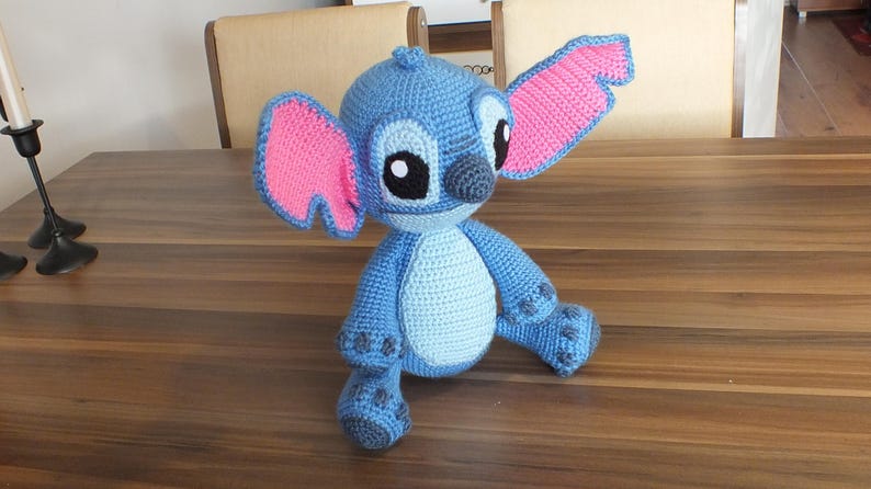 May include: A handmade blue crochet Stitch doll with pink inner ears, black eyes, and a gray nose. The plush toy is sitting on a wooden table. The doll's body is light blue, with darker blue accents on the feet and hands.