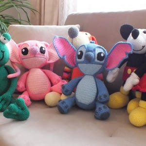 May include: A collection of handmade crochet dolls featuring popular cartoon characters. The dolls include a green frog, a pink alien, a blue alien with large ears, and Mickey and Minnie Mouse. Each doll has unique features and vibrant colors.