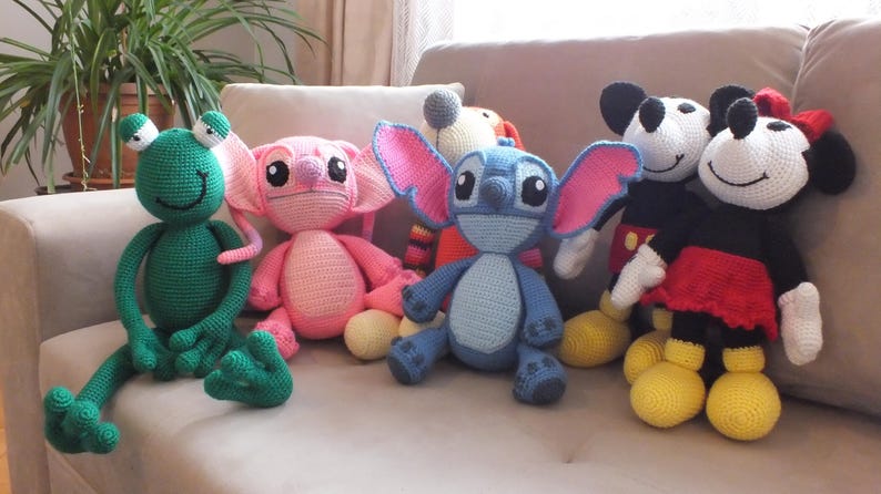 May include: A collection of handmade crochet dolls featuring popular cartoon characters. The dolls include a green frog, a pink alien, a blue alien with large ears, and Mickey and Minnie Mouse. Each doll has unique features and vibrant colors.