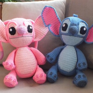 May include: Two handmade crochet dolls, one pink and one blue, sit side-by-side on a beige couch. The pink doll has large ears with pink accents, and the blue doll has large ears with pink accents. Both dolls have large black eyes.