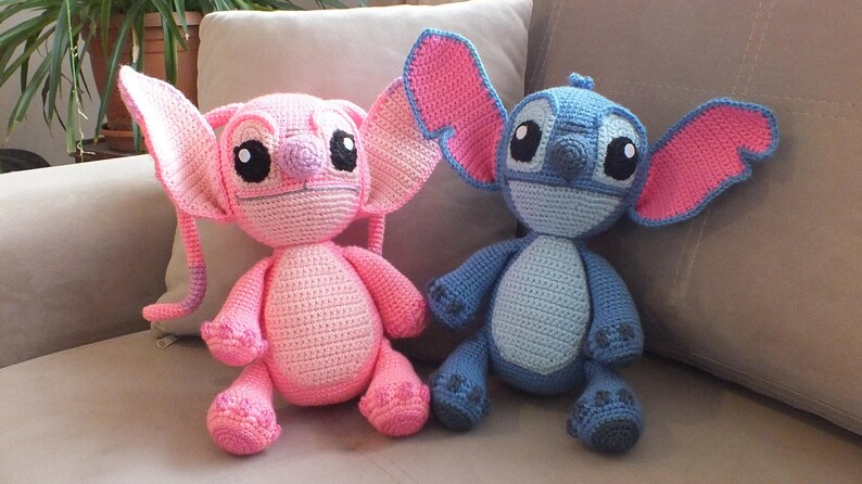 May include: Two handmade crochet dolls, one pink and one blue, sit side-by-side on a beige couch. The pink doll has large ears with pink accents, and the blue doll has large ears with pink accents. Both dolls have large black eyes.