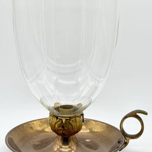 May include: A vintage candle holder with a clear glass shade and a brass base. The base features a circular dish with a handle and a decorative floral design supporting the glass shade. The candle holder is designed to hold a single candle.
