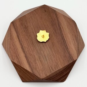 May include: A small, gold-colored flower-shaped pin is centered on a dark brown, multi-faceted wooden block. The pin has a yellow backing. The wooden block has a geometric design with angled edges and a natural wood grain.