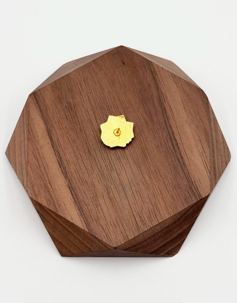 May include: A small, gold-colored flower-shaped pin is centered on a dark brown, multi-faceted wooden block. The pin has a yellow backing. The wooden block has a geometric design with angled edges and a natural wood grain.