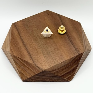 May include: A gold-colored triangle pin with a blue center and small white accents, displayed on a wooden, geometric-shaped stand. A separate gold-colored pin back is also visible. The pin is likely a piece of jewelry.