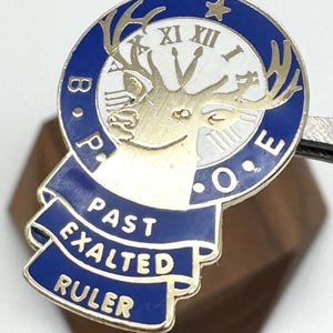 Puede incluir: Un pin dorado y azul con el texto "B.P.O.E." y "PAST EXALTED RULER". El pin presenta una esfera de reloj con n&uacute;meros romanos y una cabeza de ciervo. El pin est&aacute; sujeto por una herramienta de metal plateado.