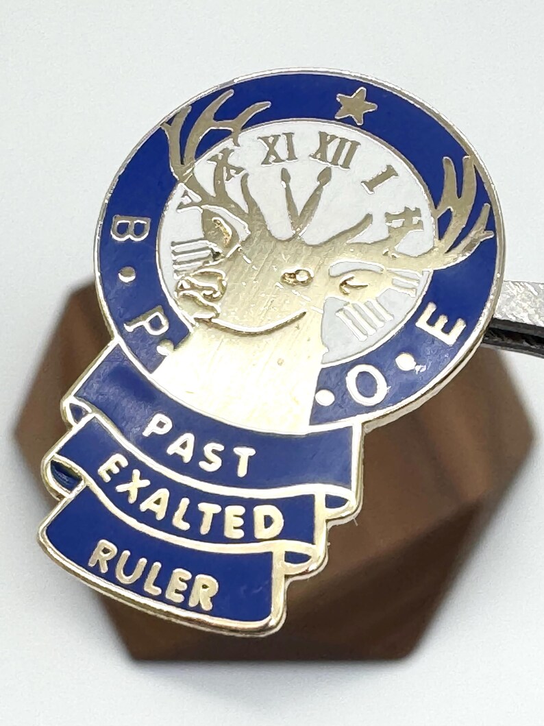 Puede incluir: Un pin dorado y azul con el texto "B.P.O.E." y "PAST EXALTED RULER". El pin presenta una esfera de reloj con n&uacute;meros romanos y una cabeza de ciervo. El pin est&aacute; sujeto por una herramienta de metal plateado.