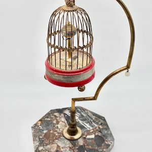 May include: A decorative birdcage clock featuring a brass cage with a small bird inside, suspended from a curved brass arm. The base is an octagonal piece of multi-colored marble. The clock face is located inside the cage, showing numbers.
