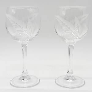 May include: Two clear glass wine glasses featuring a delicate etched fern leaf pattern on the bowl. Each glass has a slender stem and a circular base, showcasing a transparent design with frosted white leaf details on the exterior surface.