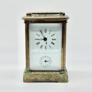 May include: A rectangular brass carriage clock with a white face featuring black Roman numerals and a smaller secondary dial with Arabic numerals. The clock has a metallic handle on top and an ornate, engraved base with intricate floral patterns.