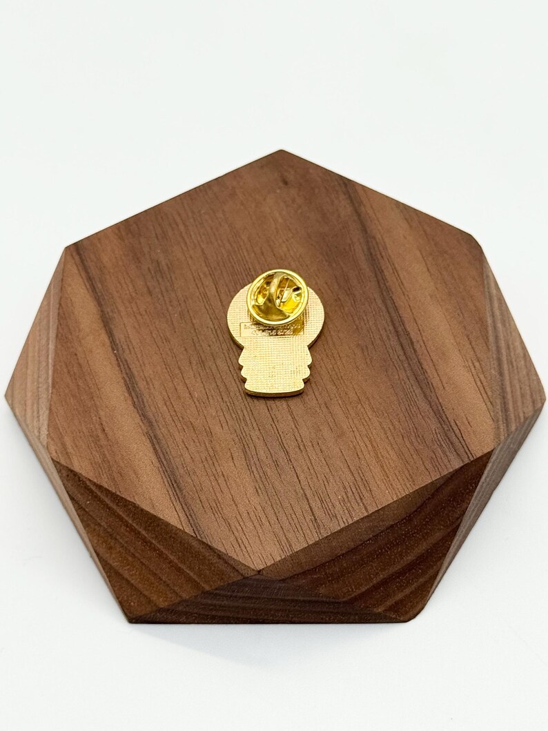Puede incluir: Un pin de esmalte dorado con un dise&ntilde;o de hongo, visto desde atr&aacute;s, mostrando el cierre del pin. El pin est&aacute; colocado sobre un soporte de madera geom&eacute;trico marr&oacute;n oscuro. El pin es probablemente un objeto de colecci&oacute;n o un accesorio.