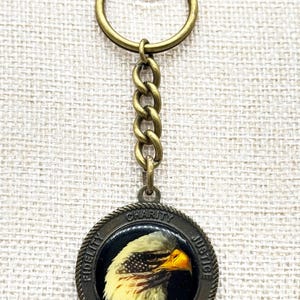 May include: A bronze-toned keychain featuring a circular pendant with an eagle's head. The pendant has the words "FIDELITY," "CHARITY," "JUSTICE," and "BROTHERLY LOVE" around the edge. The eagle's head is in shades of yellow, white, and brown.