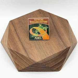 May include: A rectangular pin with the text "NETWORK WIRELESS SYSTEMS" at the top. The pin features a green and orange graphic with the AT&T logo at the bottom. The pin is set on a wooden, geometric display.