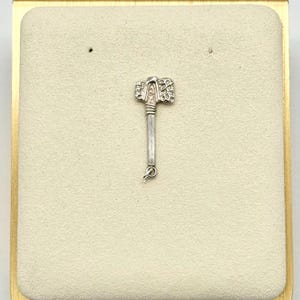 May include: A silver-colored metal charm shaped like a battle axe with a textured, patterned head and a smooth, straight handle. The charm features a small circular jump ring attached to the bottom of the handle, designed for hanging on a chain.