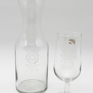 May include: Clear glass carafe and stemmed glass. The carafe has a wide base and a flared top. Both pieces have etched text that reads "ORLANDO 3 4 ESS 9-20-80" and a logo. The glass is a tulip shape.